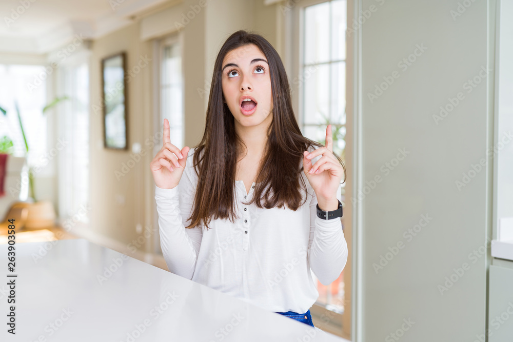 Beautiful young woman sitting on white table at home amazed and surprised looking up and pointing with fingers and raised arms.