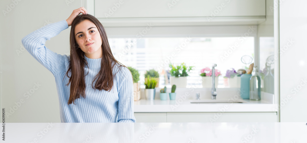 Wide angle picture of beautiful young woman sitting on white table at home confuse and wonder about question. Uncertain with doubt, thinking with hand on head. Pensive concept.