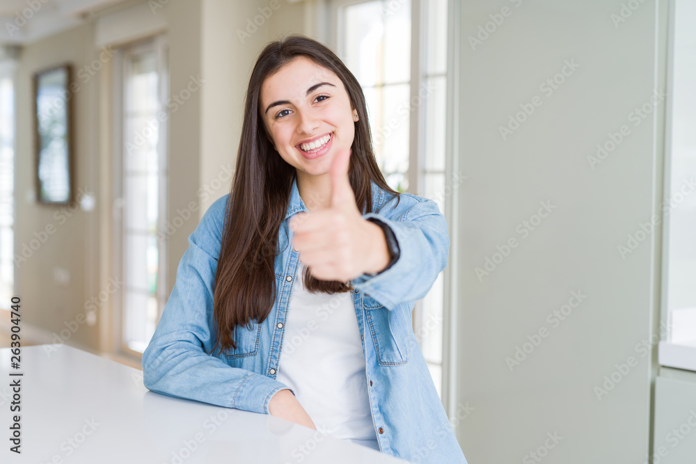 Beautiful young woman sitting on white table at home doing happy thumbs up gesture with hand. Approving expression looking at the camera with showing success.