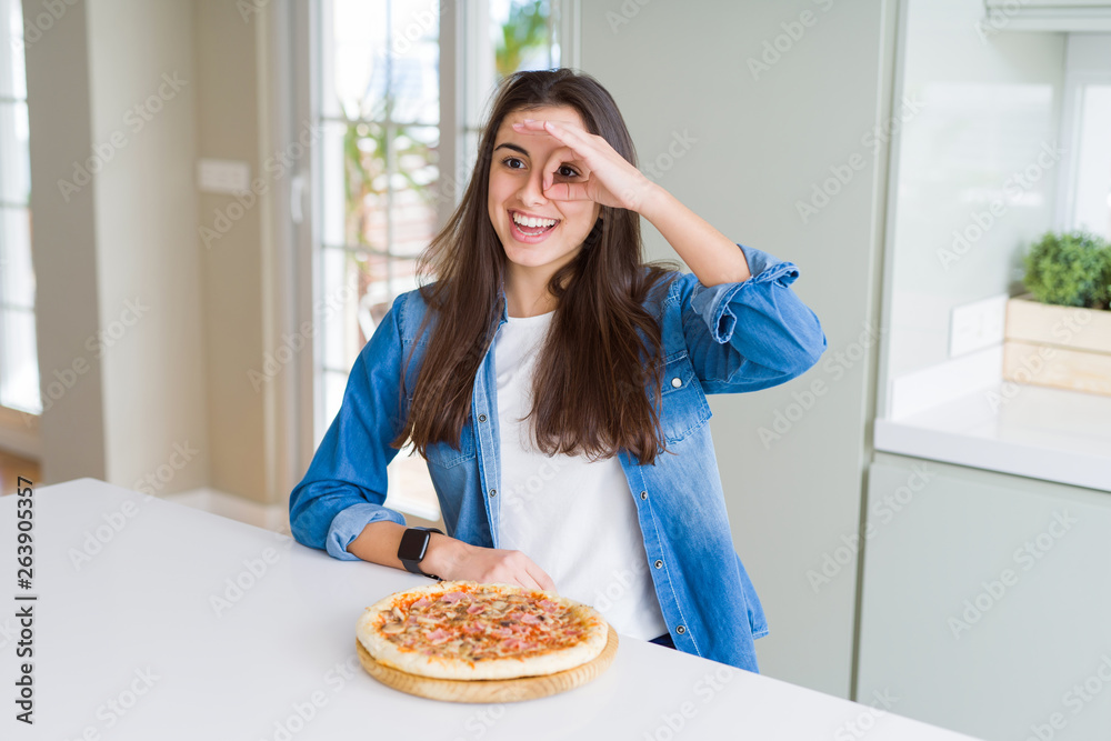 Beautiful young woman eating homemade tasty pizza at the kitchen doing ok gesture with hand smiling, eye looking through fingers with happy face.