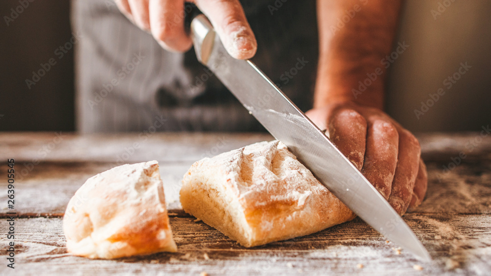 Bakery products. Homemade bread recipe. Closeup of male hands cutting fresh crusty bread with big knife on wooden table. Bakery products. Homemade bread recipe. Closeup of male hands cutting fresh crusty bread with big knife on wooden table.