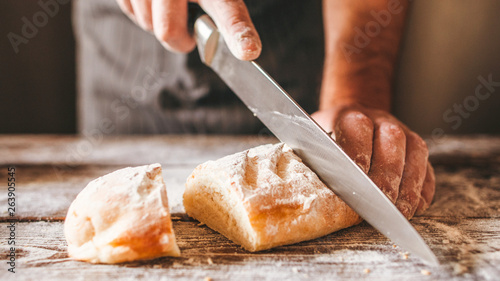 Bakery products. Homemade bread recipe. Closeup of male hands cutting fresh crusty bread with big knife on wooden table.