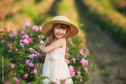 A little girl with beautiful long blond hair, dressed in a light dress and a wreath of real flowers on her head, in the garden of a tea rose