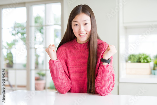 Beautiful Asian woman wearing pink sweater on white table celebrating surprised and amazed for success with arms raised and open eyes. Winner concept.