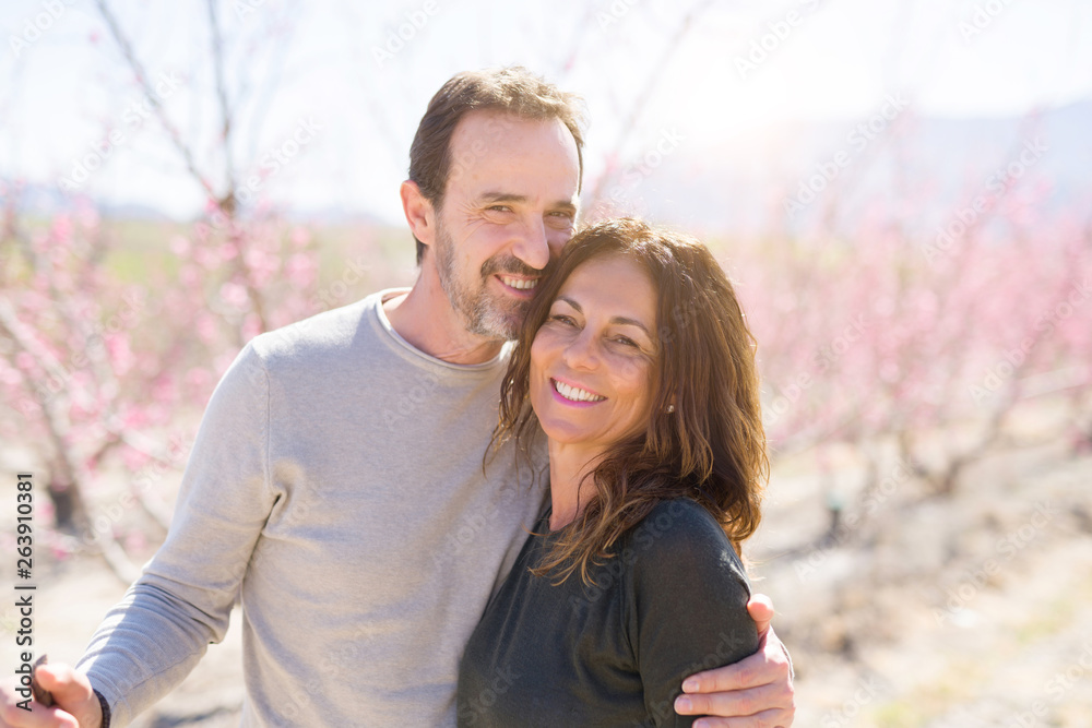 Naklejka premium Beautiful middle age senior couple smiling in love at romantic garden of peach trees with pink petals on a sunny day of spring