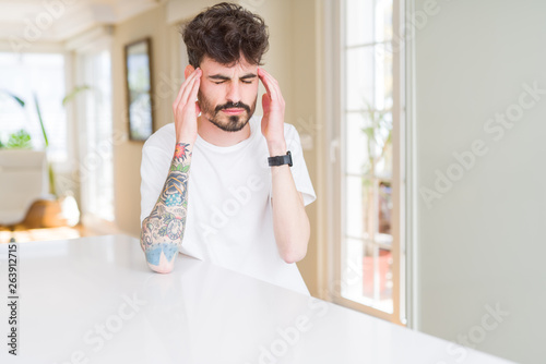 Young man wearing casual t-shirt sitting on white table with hand on head for pain in head because stress. Suffering migraine.