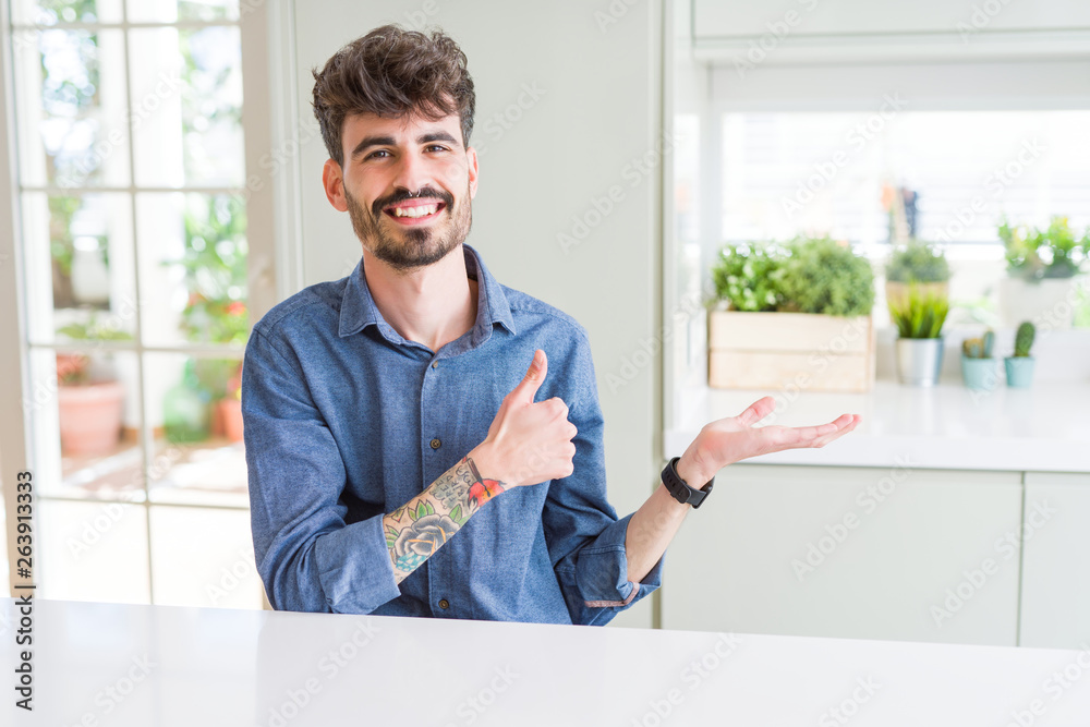 Young man wearing casual shirt sitting on white table Showing palm hand and doing ok gesture with thumbs up, smiling happy and cheerful