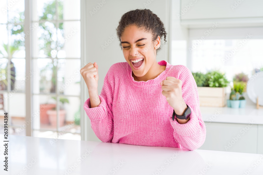 Beautiful african american woman with afro hair wearing casual pink sweater very happy and excited doing winner gesture with arms raised, smiling and screaming for success. Celebration concept.