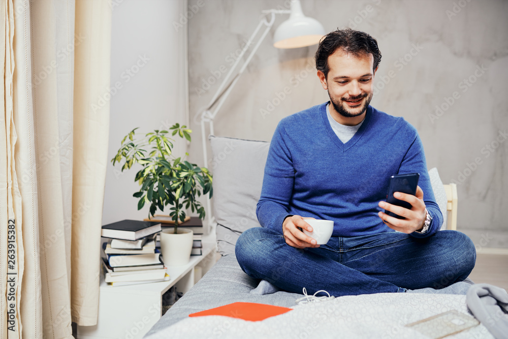 Attractive smiling arab man dressed casual sitting on bed in bedroom, drinking coffee and using smart phone for texting. Attractive smiling arab man dressed casual sitting on bed in bedroom, drinking coffee and using smart phone for texting.