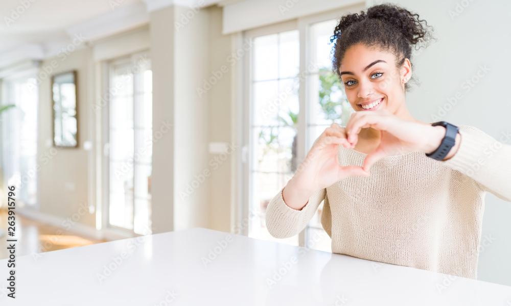 Beautiful young african american woman with afro hair sitting on table at home smiling in love showing heart symbol and shape with hands. Romantic concept. Beautiful young african american woman with afro hair sitting on table at home smiling in love showing heart symbol and shape with hands. Romantic concept.