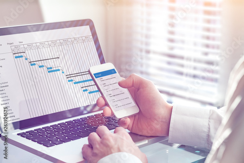 Project manager working on Gantt chart planning schedule with tasks and milestones on computer and sending email on smartphone, professional management and communication app, office interior