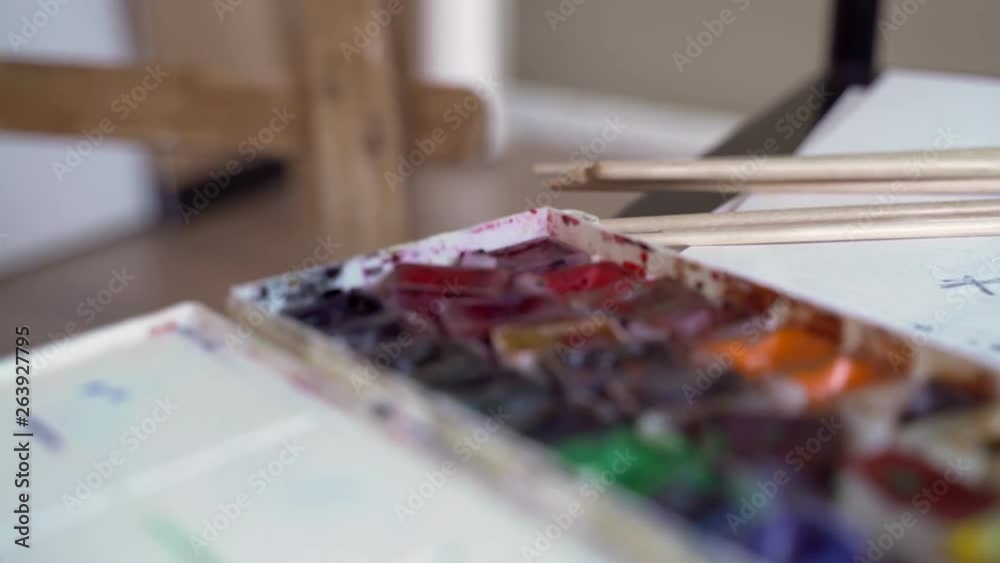 Paints are on the art table. The artist prepared brushes and a box of watercolor paints before painting. Materials for painting. Multicolored paint lie open on the table. Paints are on the art table. The artist prepared brushes and a box of watercolor paints before painting. Materials for painting. Multicolored paint lie open on the table.