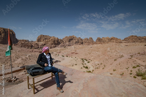 Asian man tourist sitting on wooden bench looking at rock desert landscape in Petra, Jordan from scenic viewpoint. Travel Middle East concept