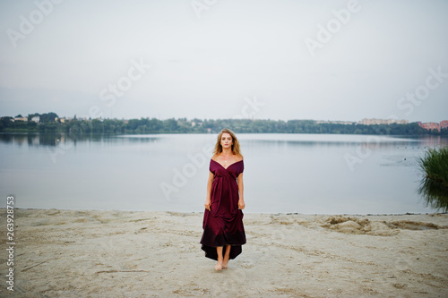 Blonde sensual barefoot woman in red marsala dress posing against lake on sand.