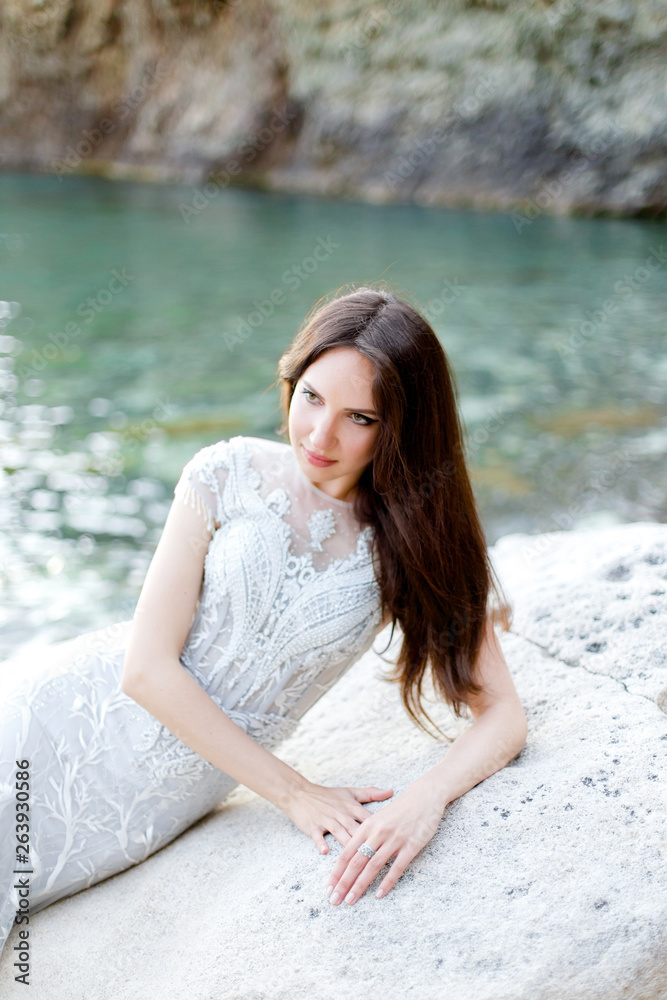 Elegant bride on a rocky beach. Young beautiful brunette woman in long light grey chiffon wedding dress embroidered with beads sitting on a rock near the sea. Full length portrait. Elegant bride on a rocky beach. Young beautiful brunette woman in long light grey chiffon wedding dress embroidered with beads sitting on a rock near the sea. Full length portrait.