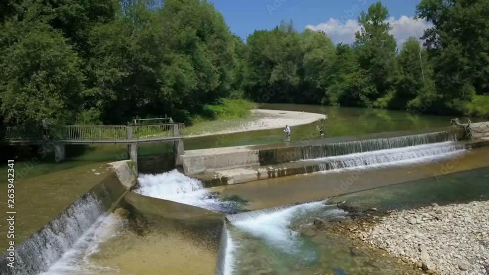 A dam was built on this river and we can see it in the front of the video. There are two fishermen fishing in the back. It's a hot summer day. Aerial shot. A dam was built on this river and we can see it in the front of the video. There are two fishermen fishing in the back. It's a hot summer day. Aerial shot.