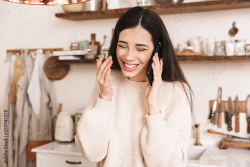 Picture of young brunette woman 30s laughing talking on smartphone at home