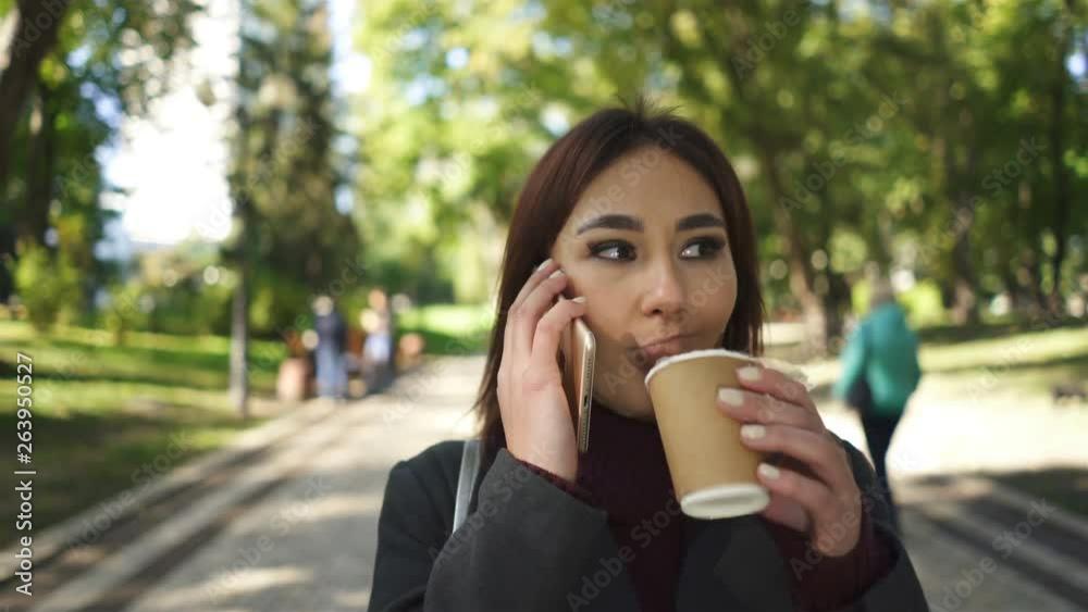   4k. Young  pretty Asian woman walk in city park with coffee and mobile. Steady shot