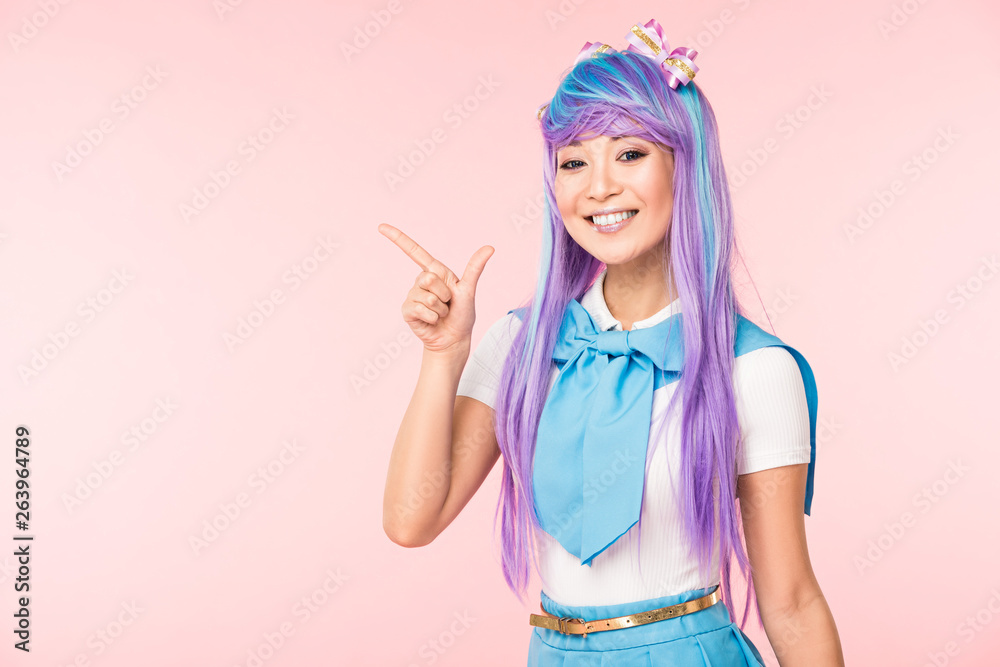 Smiling asian anime girl in purple wig pointing with finger isolated on pink Smiling asian anime girl in purple wig pointing with finger isolated on pink