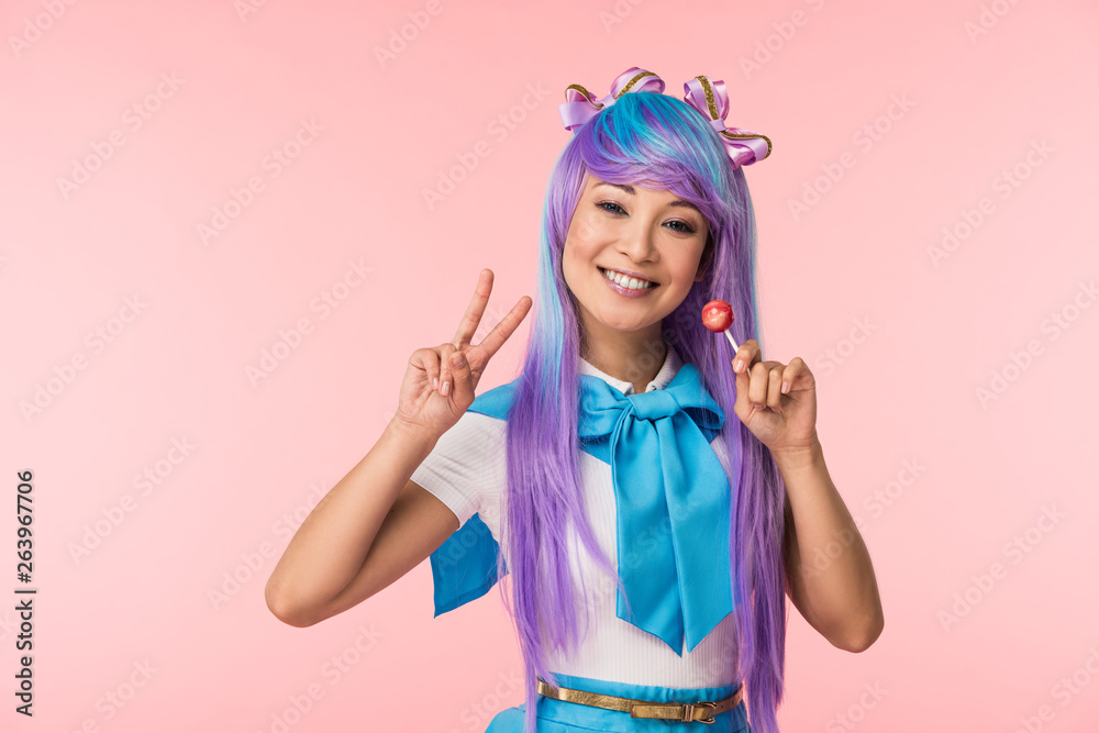 Happy asian anime girl holding lollipop and showing peace sign isolated on pink