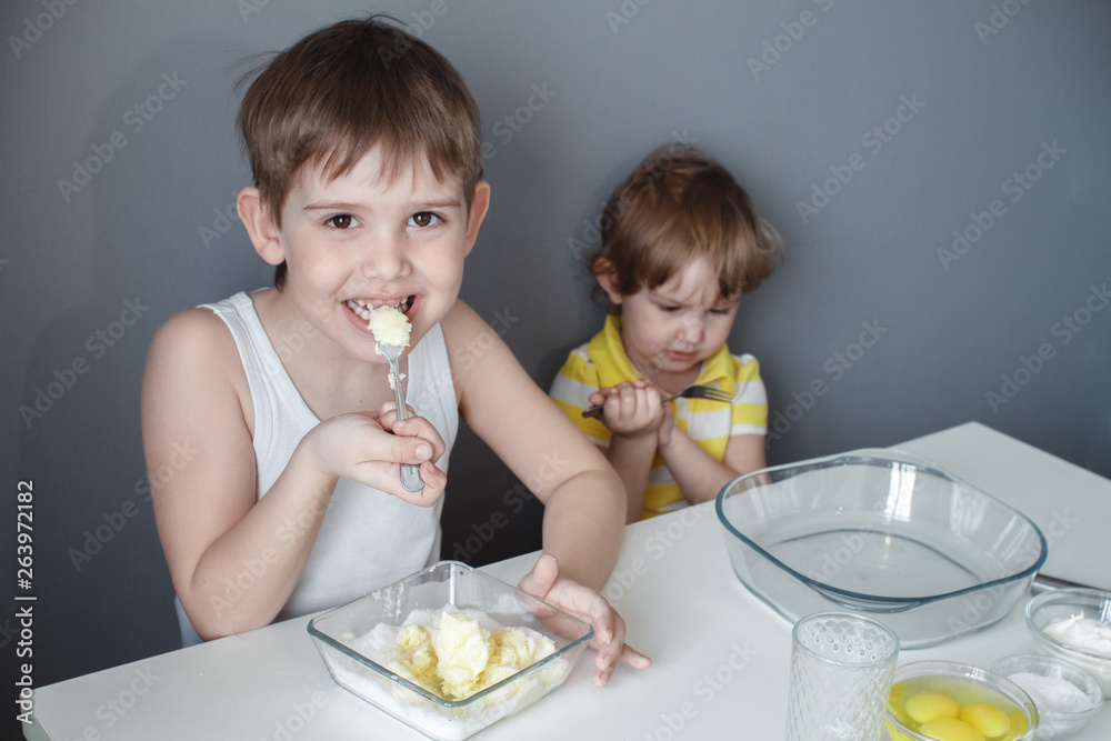 The preparation of the dessert. Two little kids are having fun making shortbread cookies, sitting at a white table and mixing ingredients. The preparation of the dessert. Two little kids are having fun making shortbread cookies, sitting at a white table and mixing ingredients.
