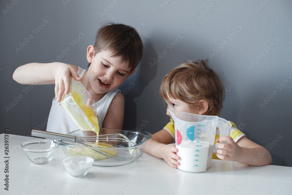 The preparation of the dessert. Two little kids are having fun making shortbread cookies, sitting at a white table and mixing ingredients. The preparation of the dessert. Two little kids are having fun making shortbread cookies, sitting at a white table and mixing ingredients.