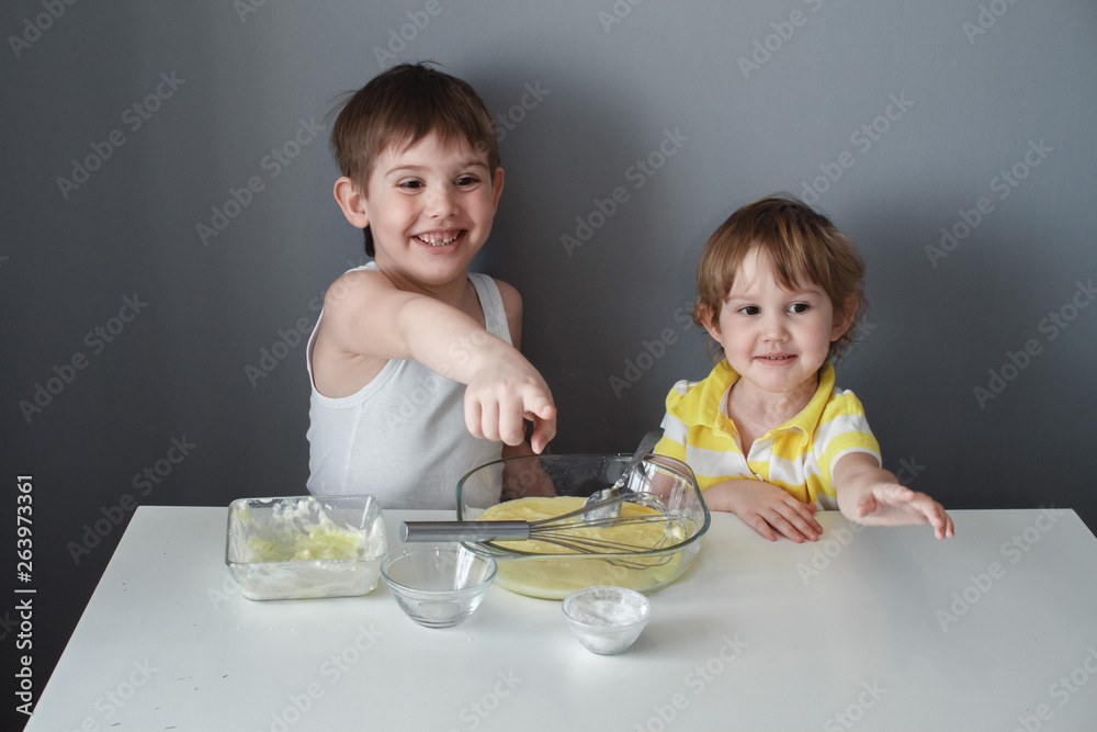 The preparation of the dessert. Two little kids are having fun making shortbread cookies, sitting at a white table and mixing ingredients The preparation of the dessert. Two little kids are having fun making shortbread cookies, sitting at a white table and mixing ingredients