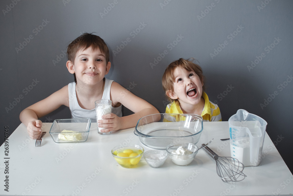 The preparation of the dessert. Two little kids are having fun making shortbread cookies, sitting at a white table and mixing ingredients. The preparation of the dessert. Two little kids are having fun making shortbread cookies, sitting at a white table and mixing ingredients.