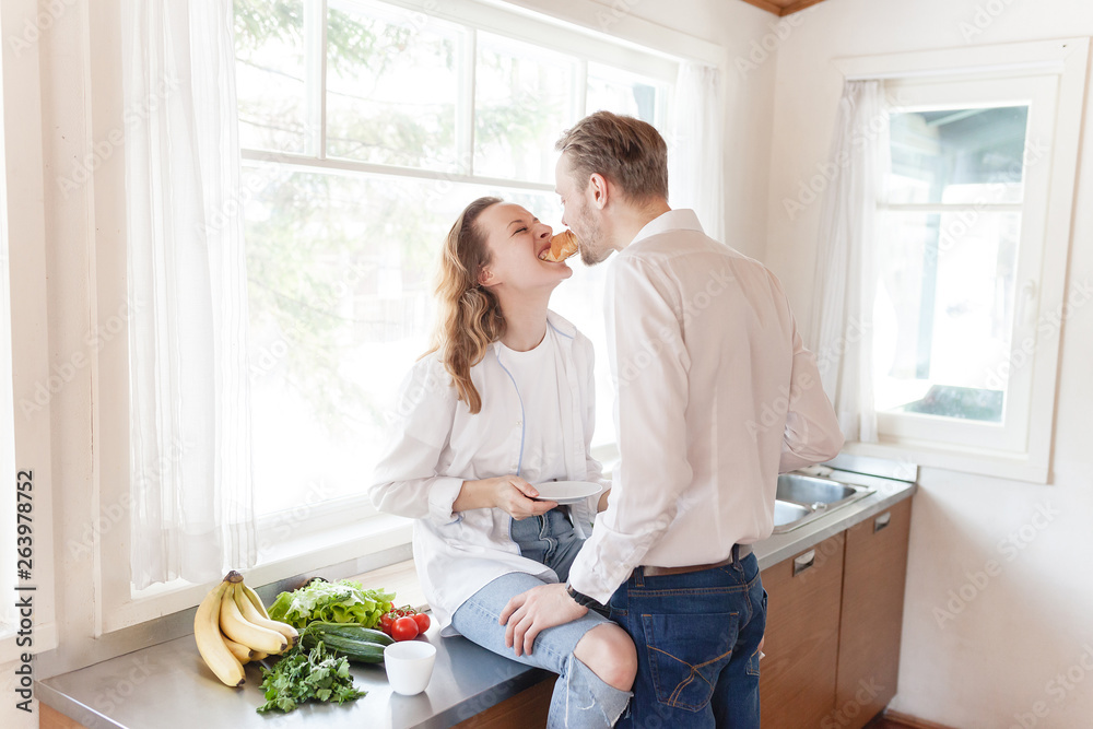 Young positive married couple is preparing lunch while sitting at his kitchen in a country house with large windows. Weekend relax concept Young positive married couple is preparing lunch while sitting at his kitchen in a country house with large windows. Weekend relax concept