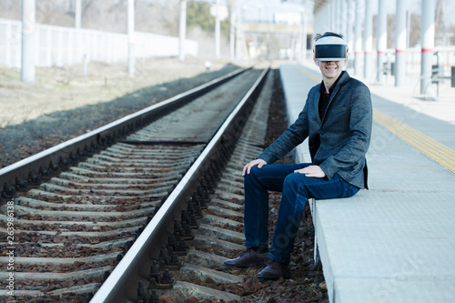 smiling young man extreme sits near the train tracks at the station wearing virtual reality glasses. Futuristic- digital technology concept. Virtual space. Vr headset and glasses