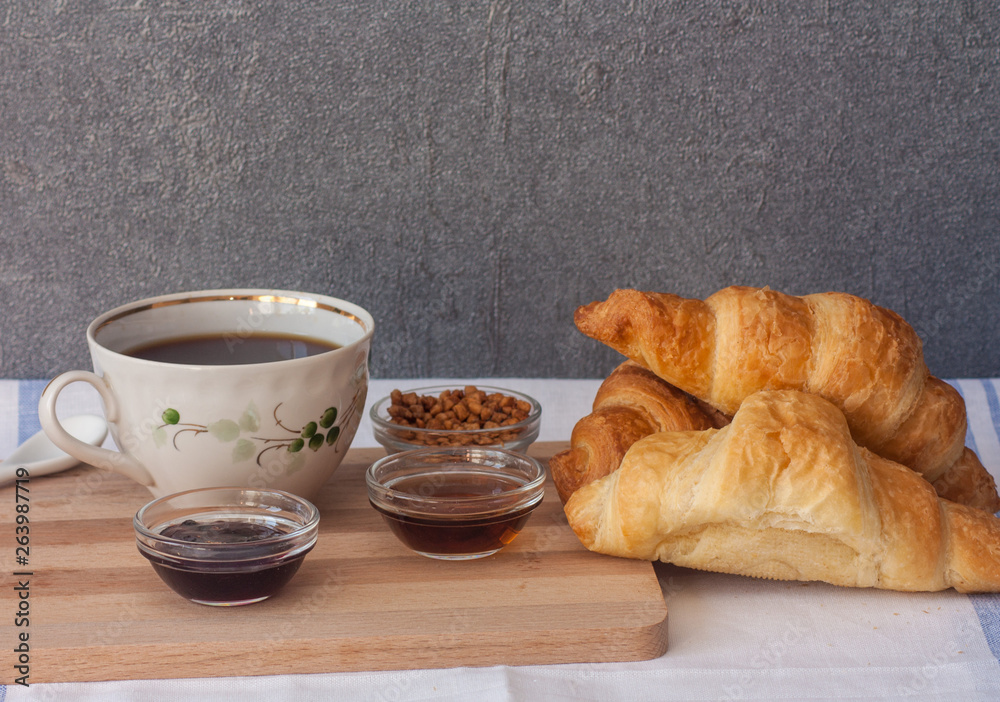 Three croissant, bowls with jam, syrup and nuts, a cup of coffee on a cutting wooden board on a light table on a background of gray wall. Three croissant, bowls with jam, syrup and nuts, a cup of coffee on a cutting wooden board on a light table on a background of gray wall.