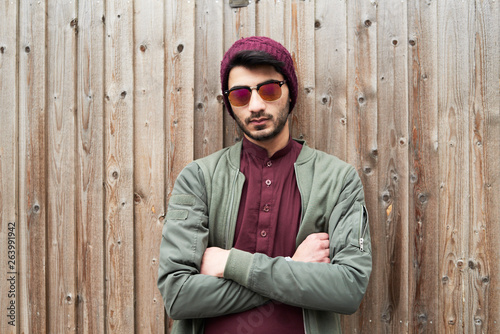 Portrait of serious bearded man in purple knit hat and shirt with arms crossed wearing sunglasses over wooden background