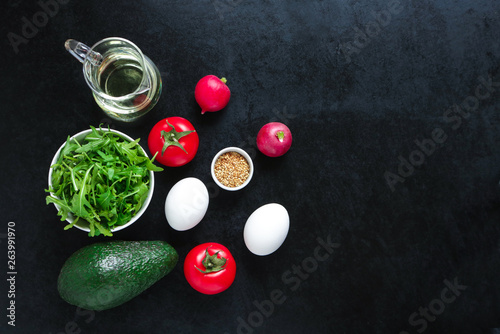 ingredients for cooking salad.boiled eggs avocado radish arugula tomatoes olive oil sesame seeds on black background top view copy space