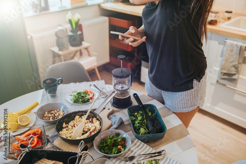 Breakfast table and woman holding smartphone in one hand navigating on social media and holding a big coffee filter glass mug with other hand  poring coffee.