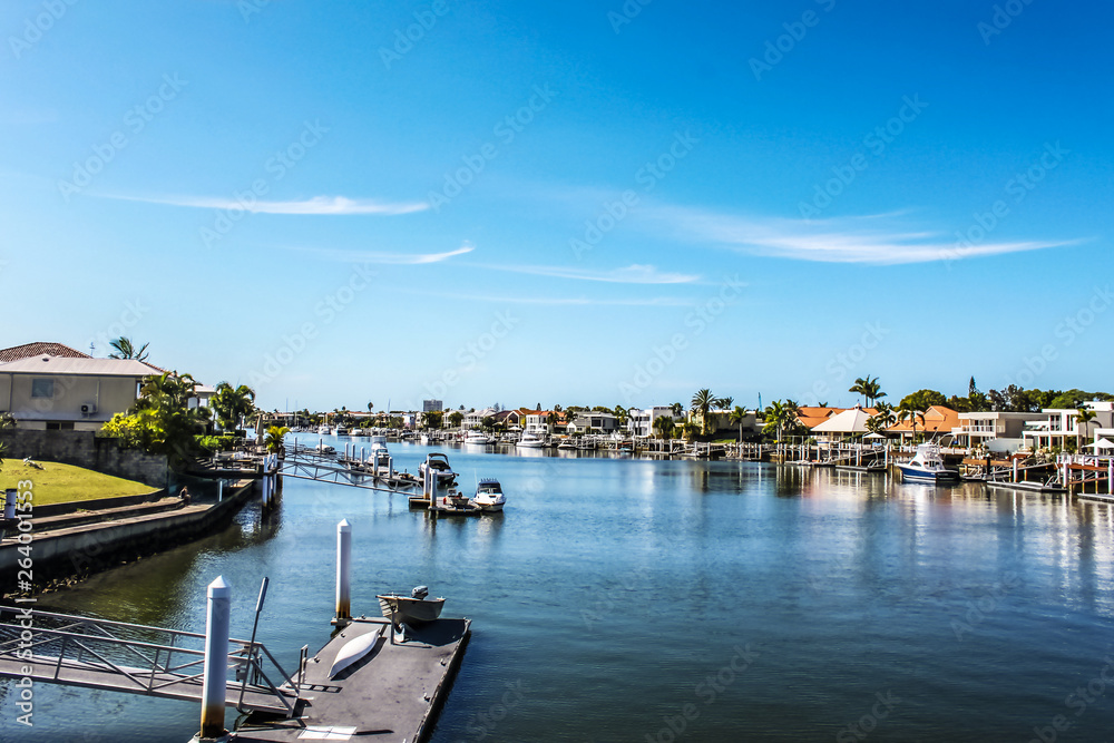 Canal in tropical setting with boat docks and moored boats by each waterside house and palm trees under blue sky