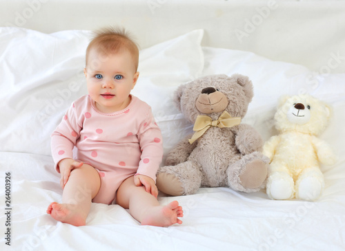 Charming blue-eyed girl 9 months on bed in pink clothes with bears toys