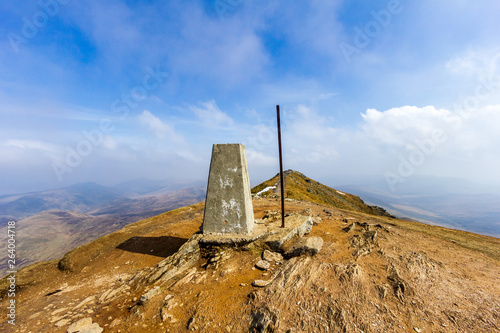 A view of the summit trig point of a Scottish mountain (Ben Vorlich) with eroded rocky soil under a majestic blue sky and altitude white clouds