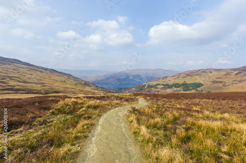 A view of a mountain valley with path,grass and lake in the background under a majestic blue sky and white clouds