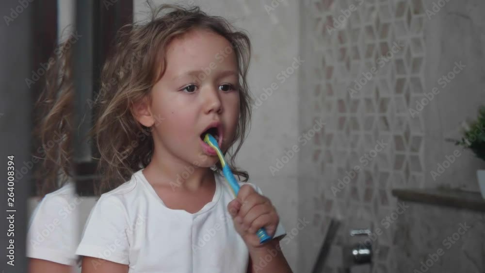 Little cute child girl in white t-shirt is brushing her teeth with toothbrush in bathroom. Portrait shot in mirror reflection. Little cute child girl in white t-shirt is brushing her teeth with toothbrush in bathroom. Portrait shot in mirror reflection.