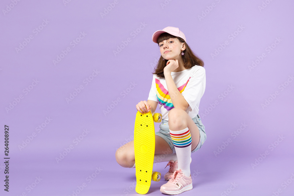 Cute teen girl in vivid clothes sitting holding yellow skateboard, put hand prop up on chin isolated on violet pastel wall background. People sincere emotions, lifestyle concept. Mock up copy space. Cute teen girl in vivid clothes sitting holding yellow skateboard, put hand prop up on chin isolated on violet pastel wall background. People sincere emotions, lifestyle concept. Mock up copy space.
