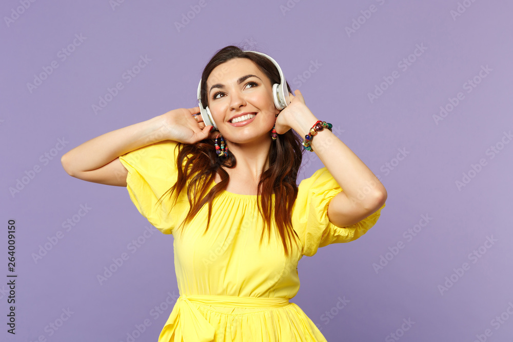 Charming cute young woman in yellow dress looking up listen music putting hands on headphones isolated on pastel violet wall background. People sincere emotions, lifestyle concept. Mock up copy space. Charming cute young woman in yellow dress looking up listen music putting hands on headphones isolated on pastel violet wall background. People sincere emotions, lifestyle concept. Mock up copy space.