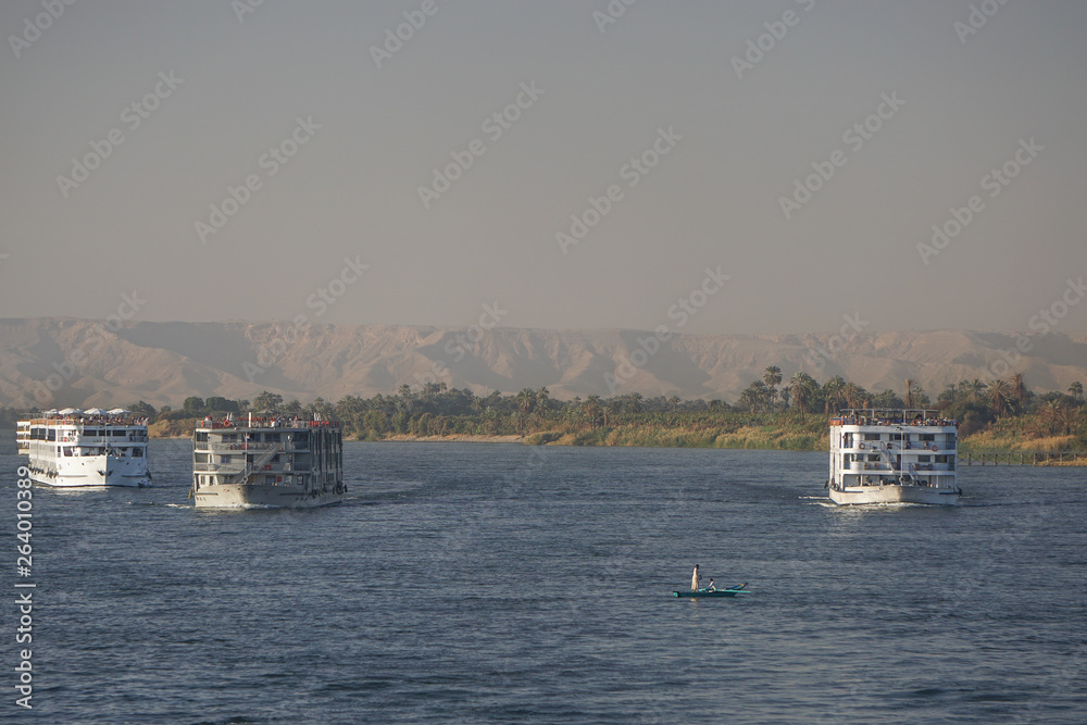 Nile River, Egypt: Two men in a rowboat watch as several river boats sail upstream on the Nile River; the dunes of the west bank in the background. Nile River, Egypt: Two men in a rowboat watch as several river boats sail upstream on the Nile River; the dunes of the west bank in the background.