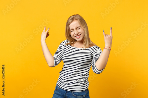 Crazy young woman keeping eyes closed, showing horns up gesture, depicting heavy metal rock sign isolated on yellow orange background. People sincere emotions, lifestyle concept. Mock up copy space.