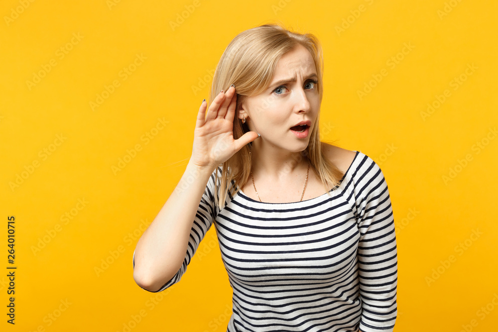 Portrait of puzzled young woman in striped clothes trying to hear you with hand near ear isolated on yellow orange background in studio. People sincere emotions, lifestyle concept. Mock up copy space. Portrait of puzzled young woman in striped clothes trying to hear you with hand near ear isolated on yellow orange background in studio. People sincere emotions, lifestyle concept. Mock up copy space.
