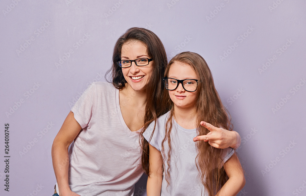 Happy fun young casual mother showing the horns sign two fingers and hugging her cute kid girl on violet wall background. Family in fashion eye glasses. Happy fun young casual mother showing the horns sign two fingers and hugging her cute kid girl on violet wall background. Family in fashion eye glasses.