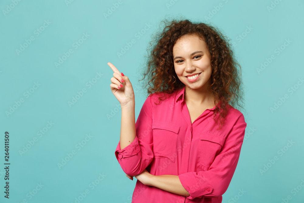 Portrait of smiling african girl in pink casual clothes pointing index finger aside isolated on blue turquoise wall background in studio. People sincere emotions lifestyle concept. Mock up copy space. Portrait of smiling african girl in pink casual clothes pointing index finger aside isolated on blue turquoise wall background in studio. People sincere emotions lifestyle concept. Mock up copy space.