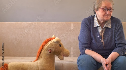 Elderly woman sits and plays with her grandson on couch. Child loves grandmother very much. Boy kisses and hugs his beloved grandmother. Kid shows how he can use computer tablet.