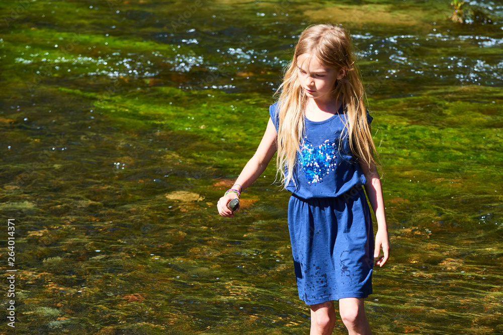 Child cute blond girl playing in the creek. Girl walking in forest stream, looking for pebbles and exploring nature. Summer children fun. Children summer activities Child cute blond girl playing in the creek. Girl walking in forest stream, looking for pebbles and exploring nature. Summer children fun. Children summer activities