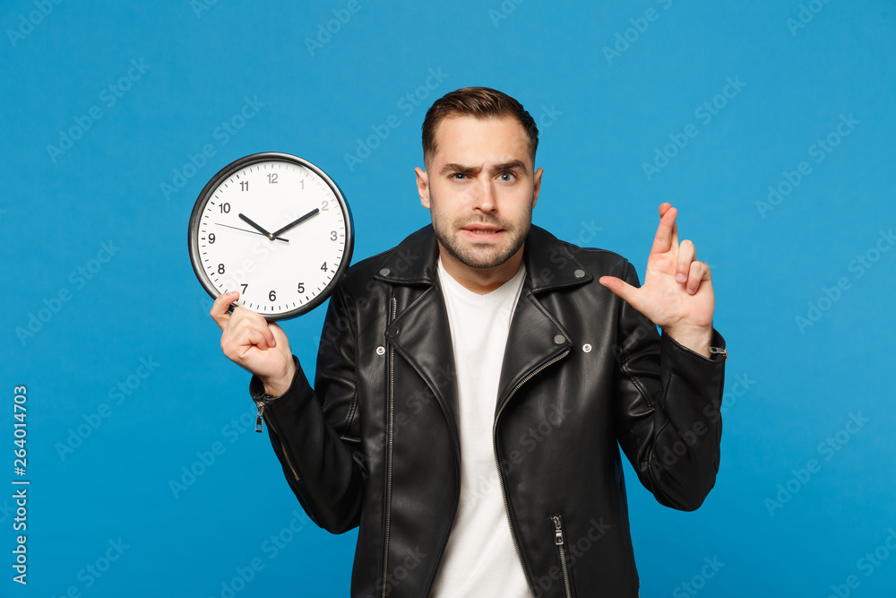 Sad upset stylish young unshaven man in black leather jacket white t-shirt holding round clock isolated on blue wall background studio portrait. People lifestyle concept. Hurry up. Mock up copy space. Sad upset stylish young unshaven man in black leather jacket white t-shirt holding round clock isolated on blue wall background studio portrait. People lifestyle concept. Hurry up. Mock up copy space.