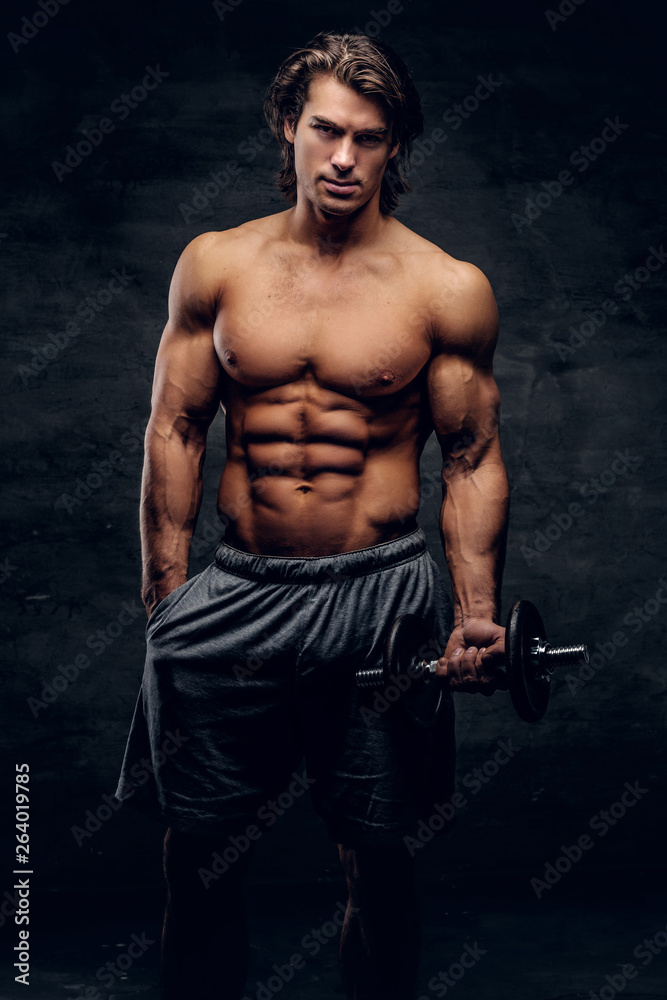 Beautiful young bodybuilder is posing with dumbbell. He has naked torso.